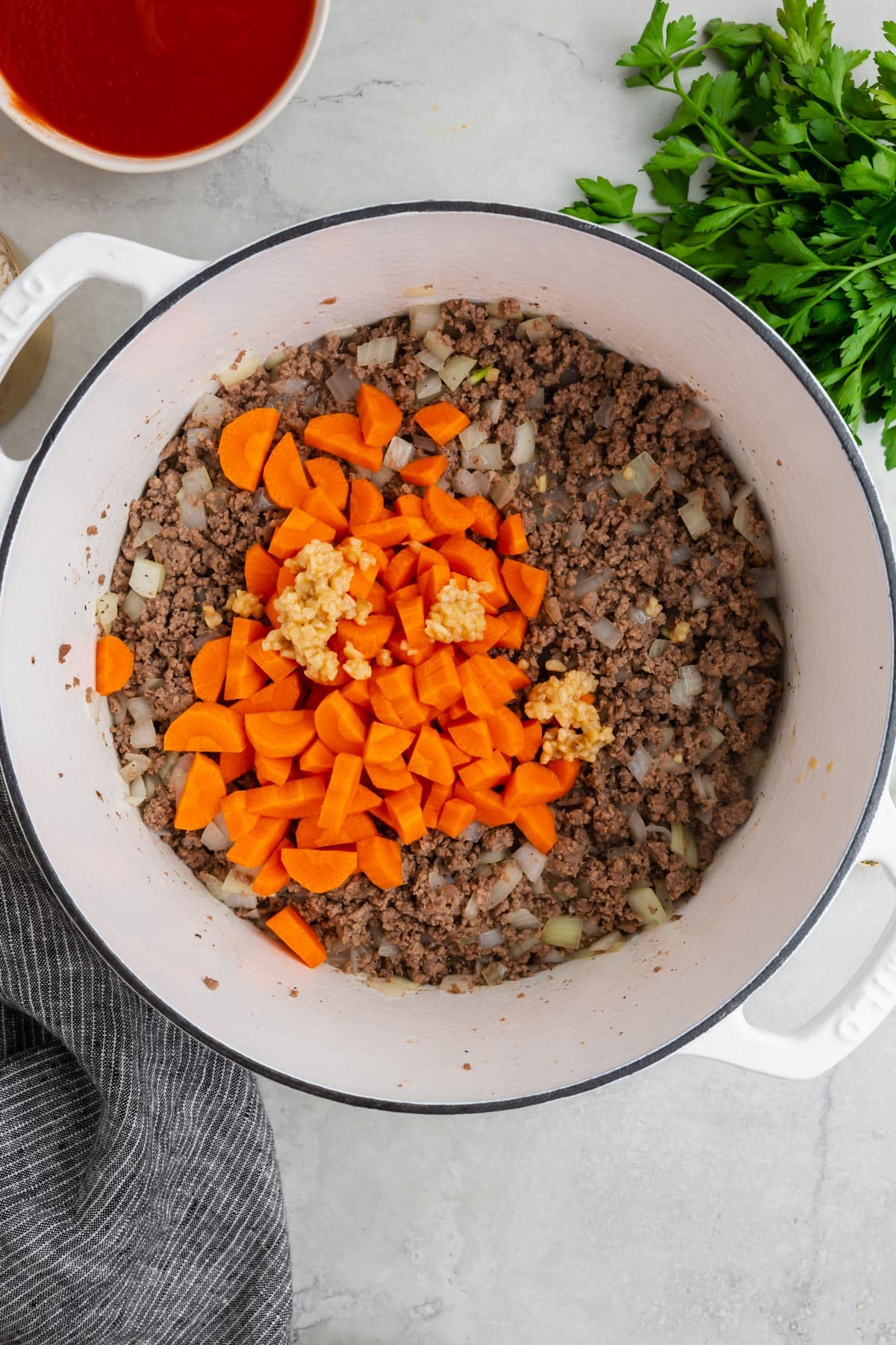 An overhead view of a white dutch oven with ground beef topped with diced carrots and minced garlic.