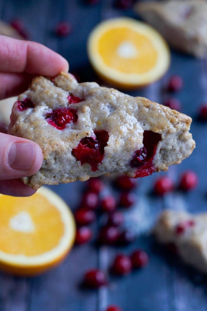 A hand holding a vegan cranberry scone with orange halves and fresh cranberries in the background.