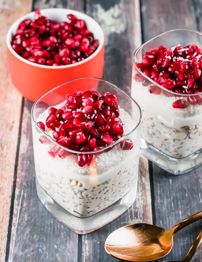 Two glasses of overnight oats topped with coconut shreds and pomegranate arils in front of a bowl of pomegranate arils.