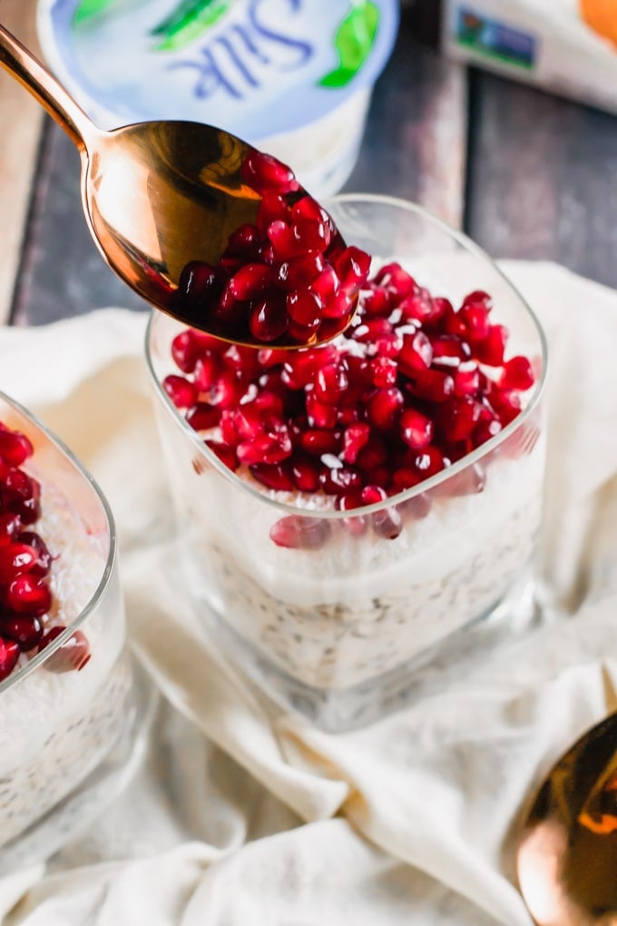 A spoon placing pomegranate arils on top of a glass of overnight oats.