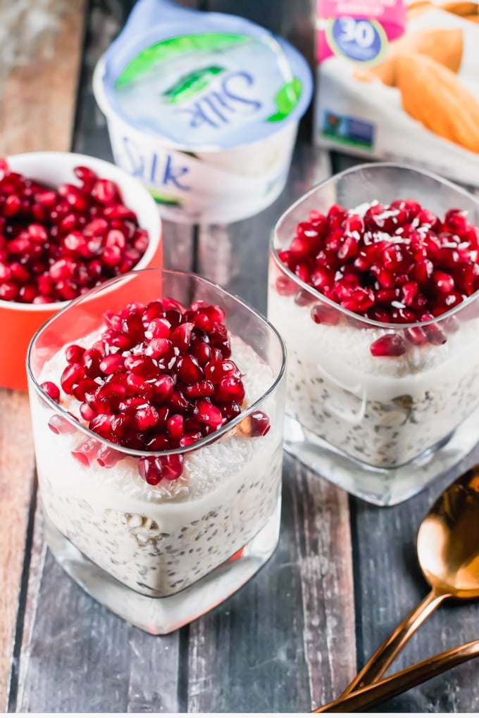 Two glasses of overnight oats with pomegranate on top in front of a yogurt cup and a bowl of pomegranate arils.
