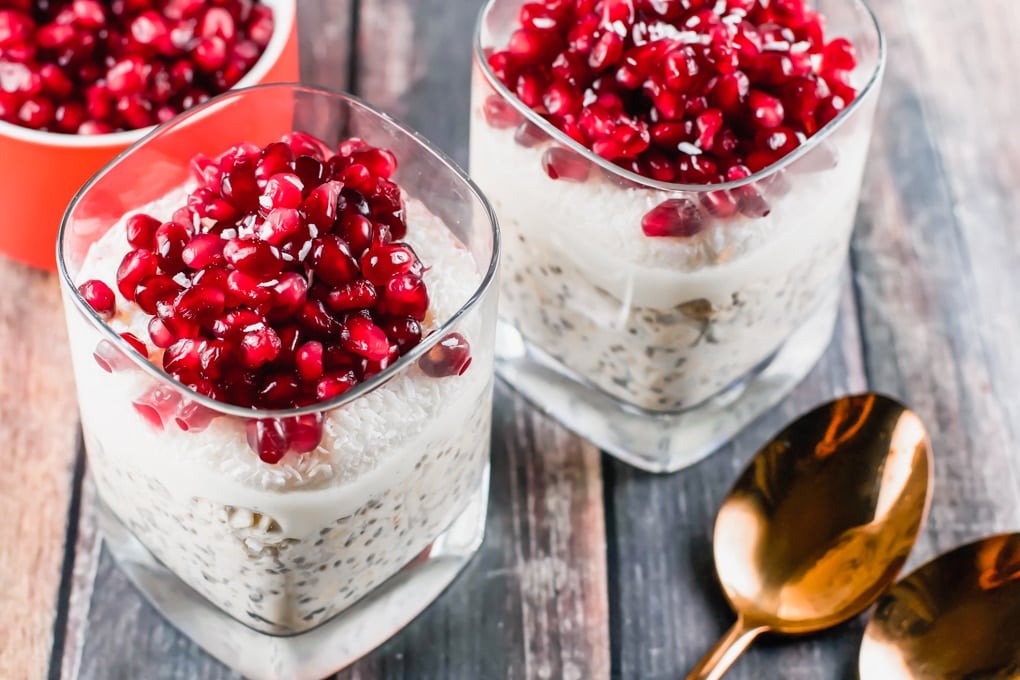 Two glasses of overnight oats topped with coconut and pomegranate arils in front of a bowl of pomegranate arils.