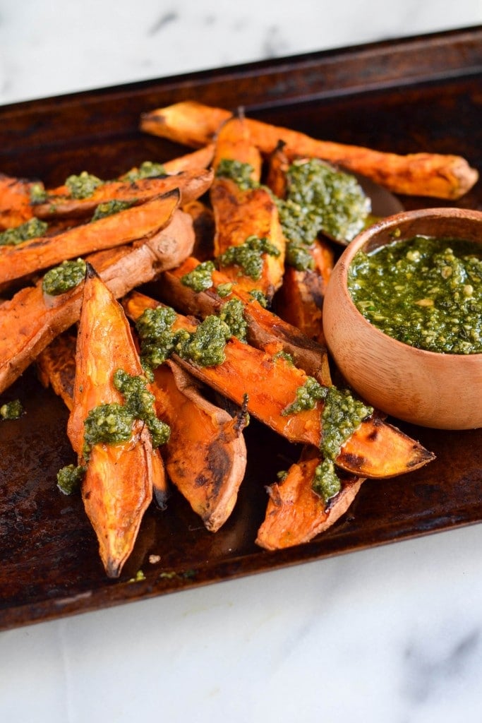 Sweet potato wedges topped with parsley pesto on a baking sheet with a bowl of pesto on the side.