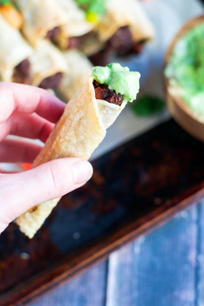 A hand holding a black bean taquito topped with avocado dip. 