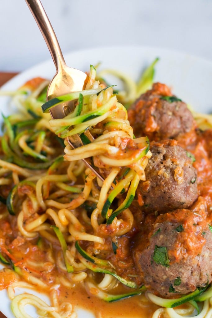 A fork twirling zucchini noodles in a roasted red pepper sauce with some turkey meatballs.