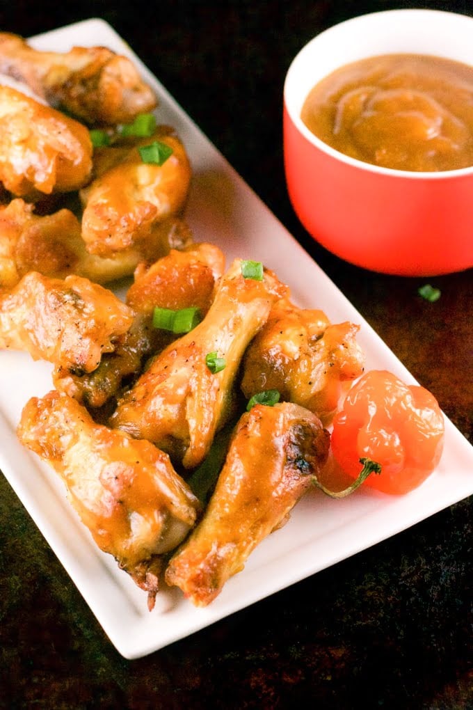 A white rectangular plate of baked mango habanero chicken wings with a bowl of mango habanero sauce in the background.