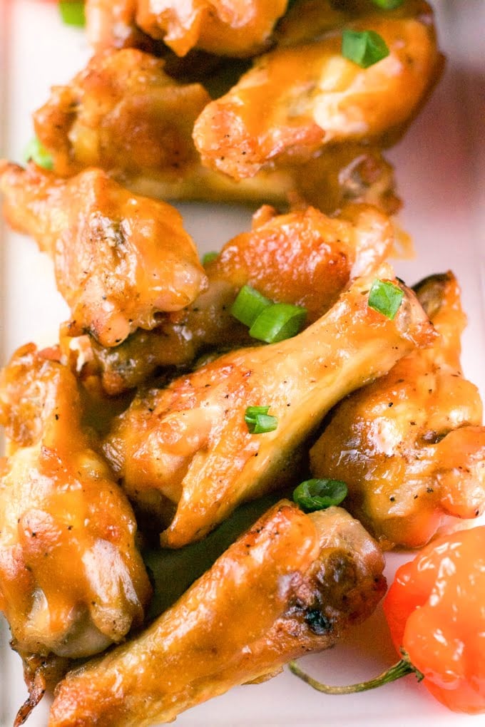 A pile of mango habanero chicken wings on a platter topped with green onions with habanero peppers on the side. 