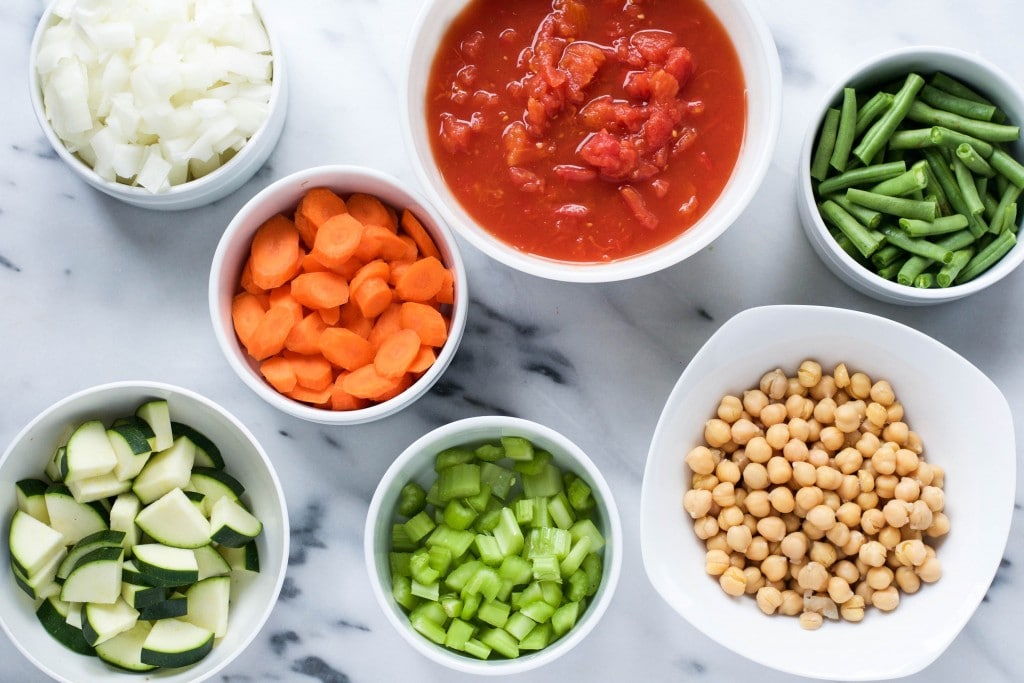Bowls of celery, zucchini, green beans, chickpeas, stewed tomatoes, carrots, and yellow onion to make minestrone soup. 