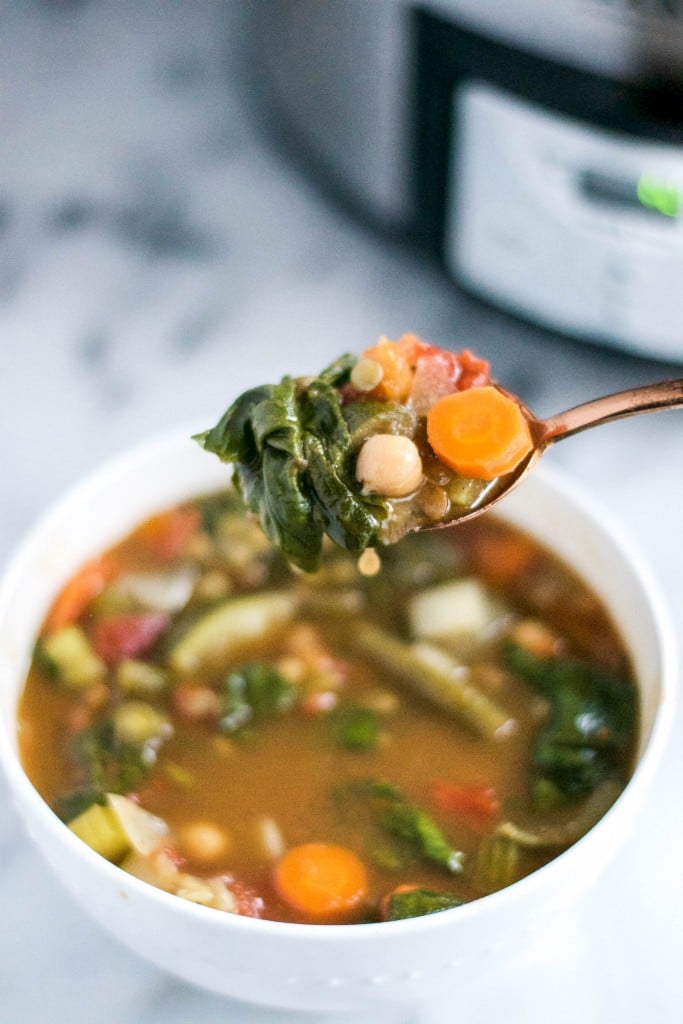 A spoon of vegetable chickpea soup over a bowl of soup with a slow cooker in the background.