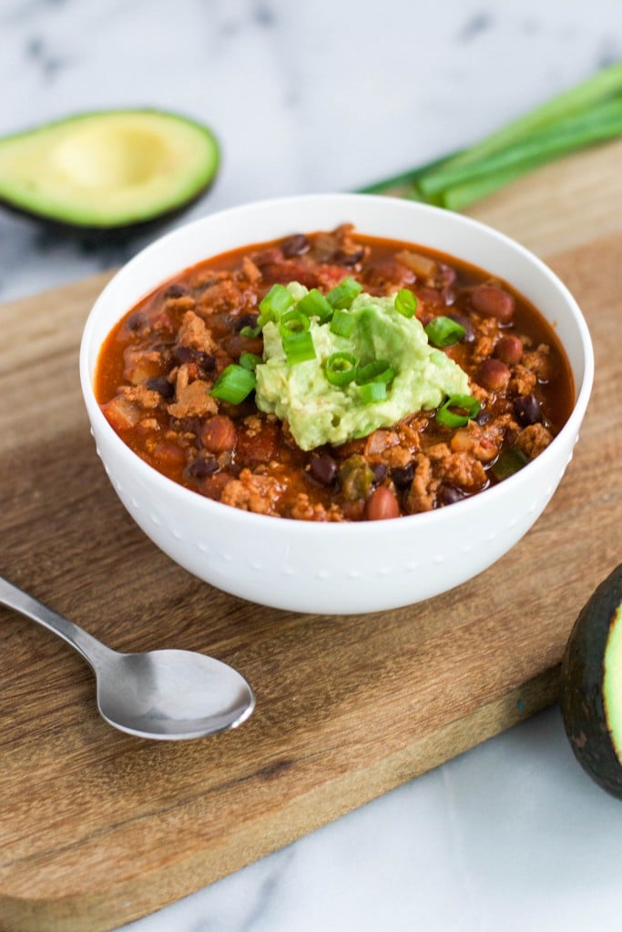 A bowl of turkey chili topped with guacamole and green onions sitting on a wooden board.