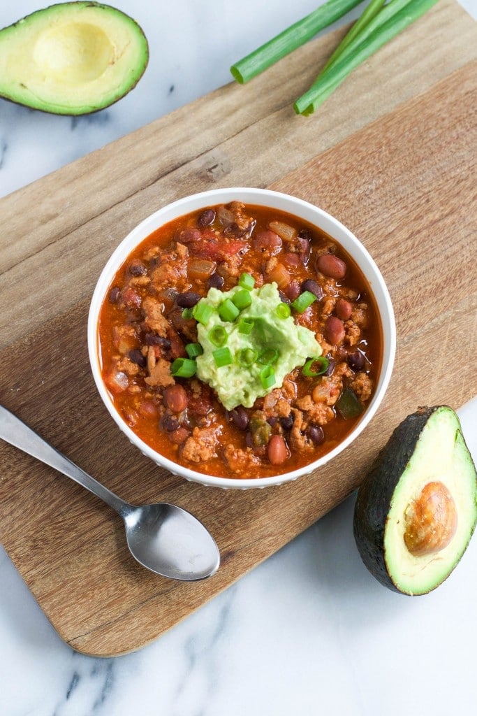 An overhead view of a bowl of turkey chili topped with guacamole and green onions on a wooden board with a spoon.
