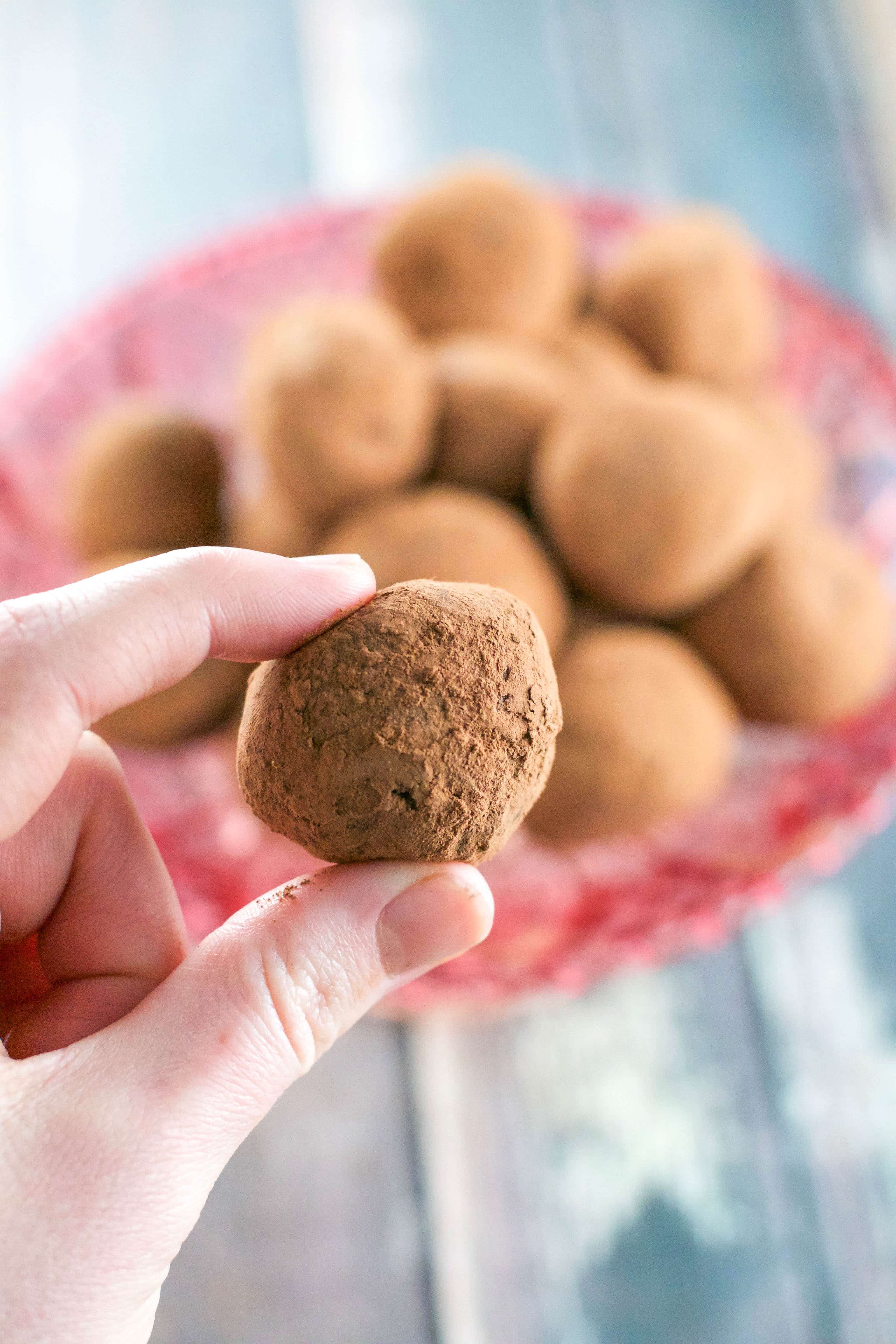 A hand holding a single vegan chocolate truffle with more truffles in the background on a pink cake stand.