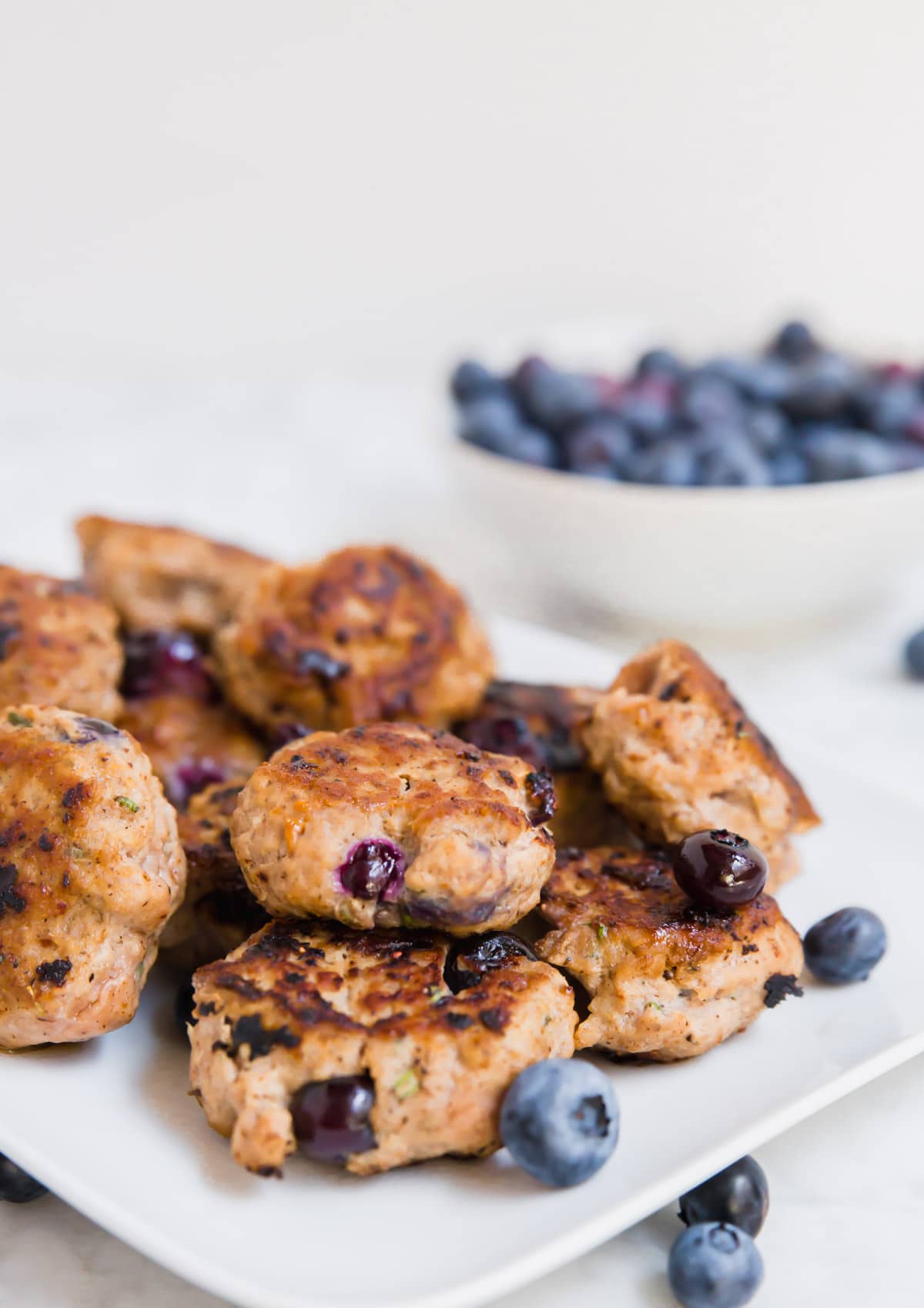 A photo of a white plate with cooked blueberry turkey breakfast sausage with a bowl of fresh blueberries in the background. 