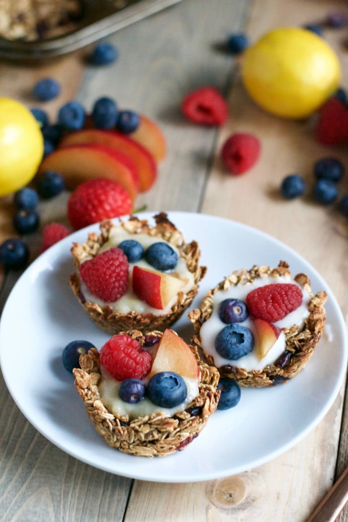 Three vegan granola cups filled with yogurt, raspberries, blueberries and plums with fresh fruit and lemons in the background.