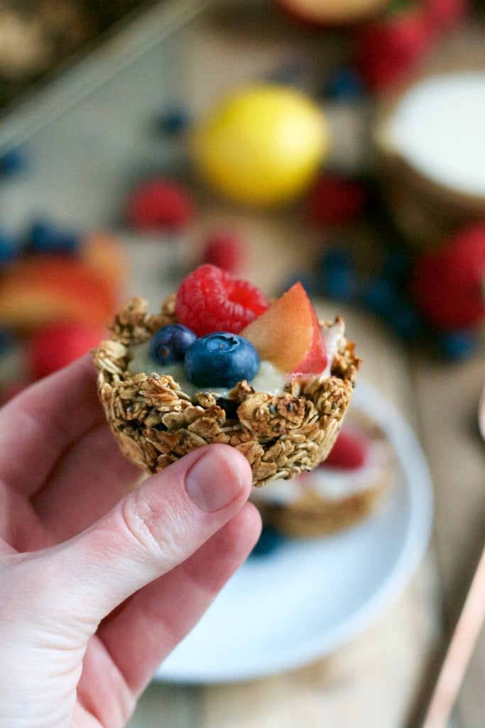 A hand holding a vegan granola cup filled with yogurt, blueberries, raspberries and plums. 