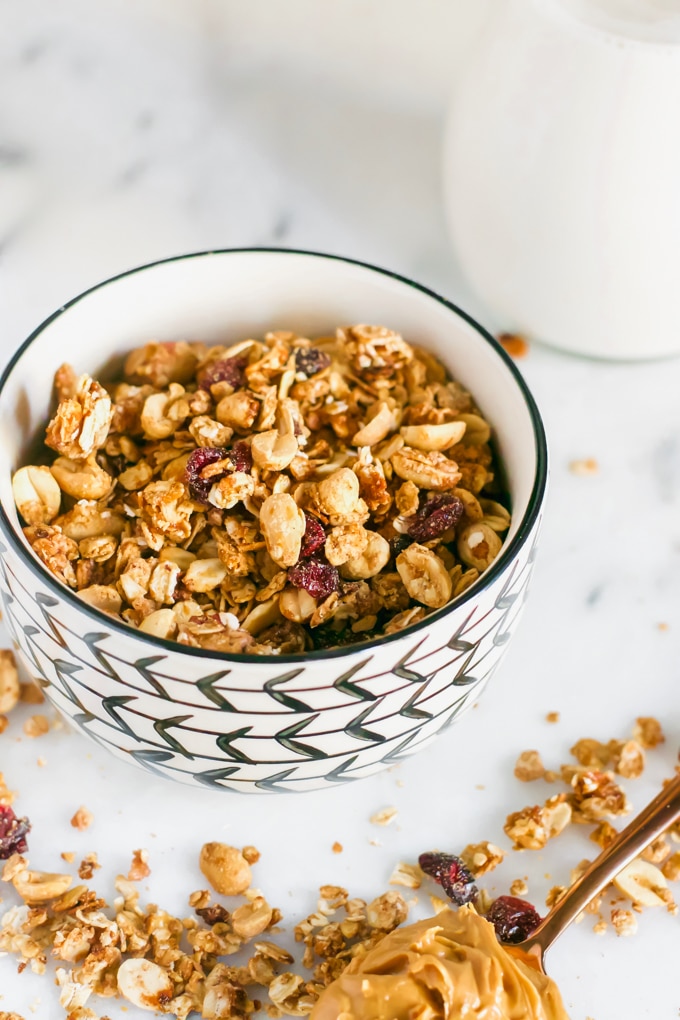 A bowl of granola topped with dried cranberries with a glass of milk in the background and a spoon of peanut butter in the foreground.