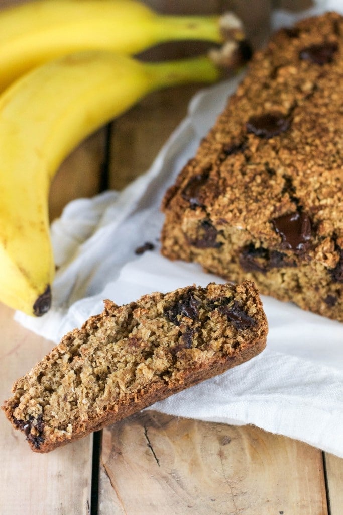 A slice of vegan banana chocolate chunk bread in front of a loaf of bread and a bunch of bananas. 