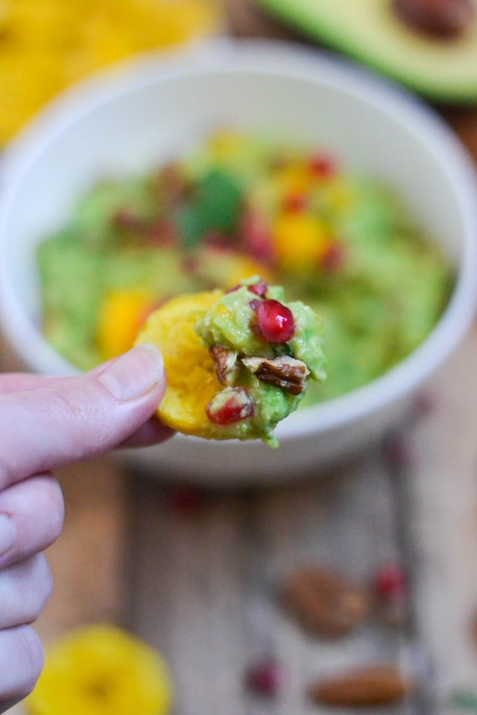A hand holding a plantain chip topped with guacamole, pomegranate aril and pecans.