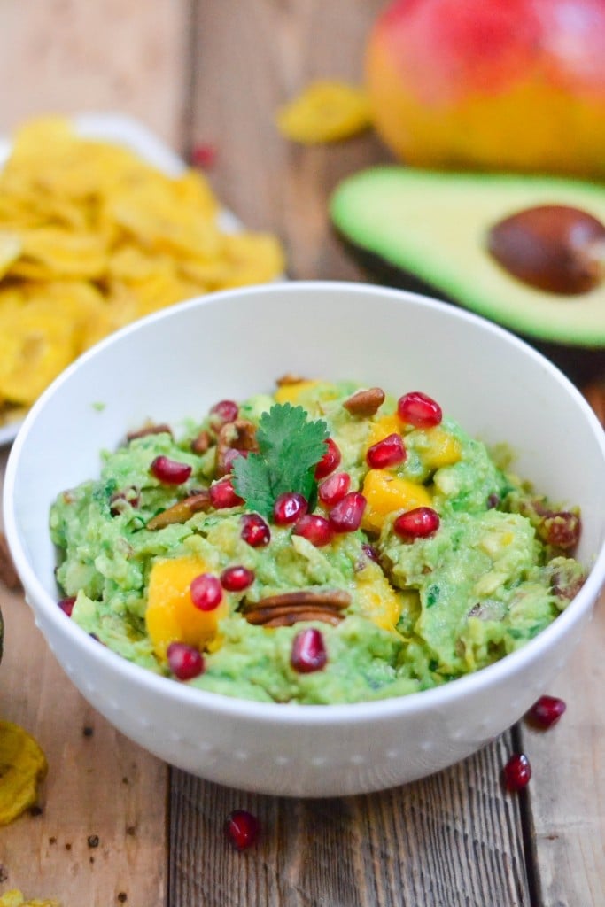 A bowl of guacamole topped with mango, pomegranate arils, and pecans in front of a tray of plantain chips and an avocado half. 