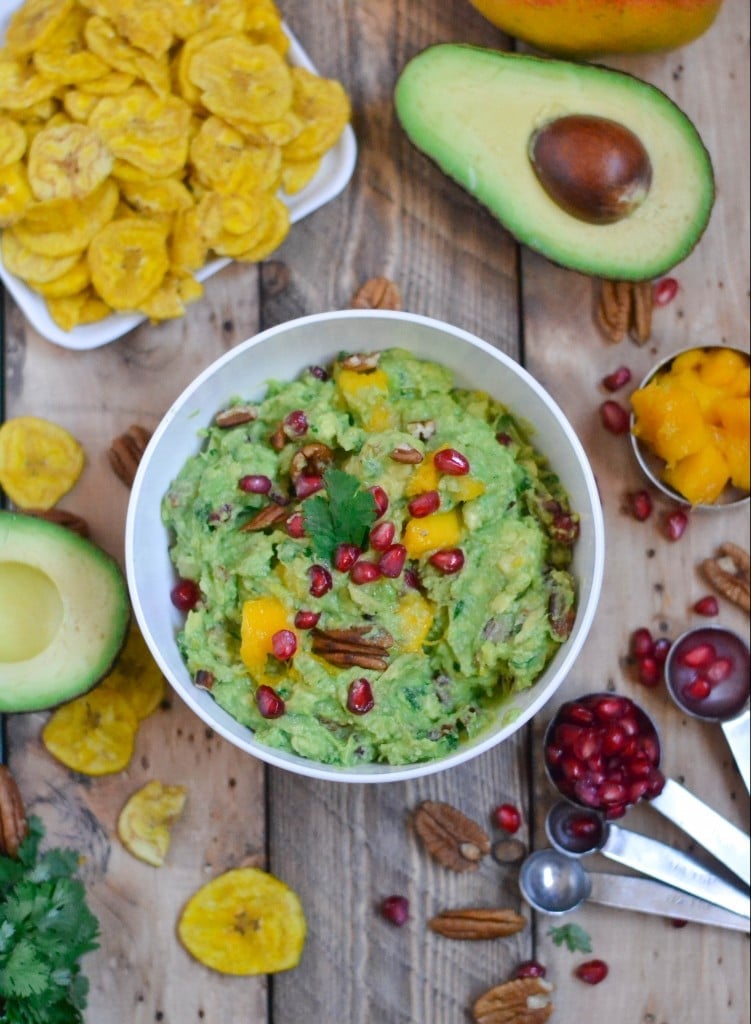 A bowl of guacamole topped with pecans, mango, and pomegranate arils with a side of plantain chips, an avocado half and fresh pomegranate.
