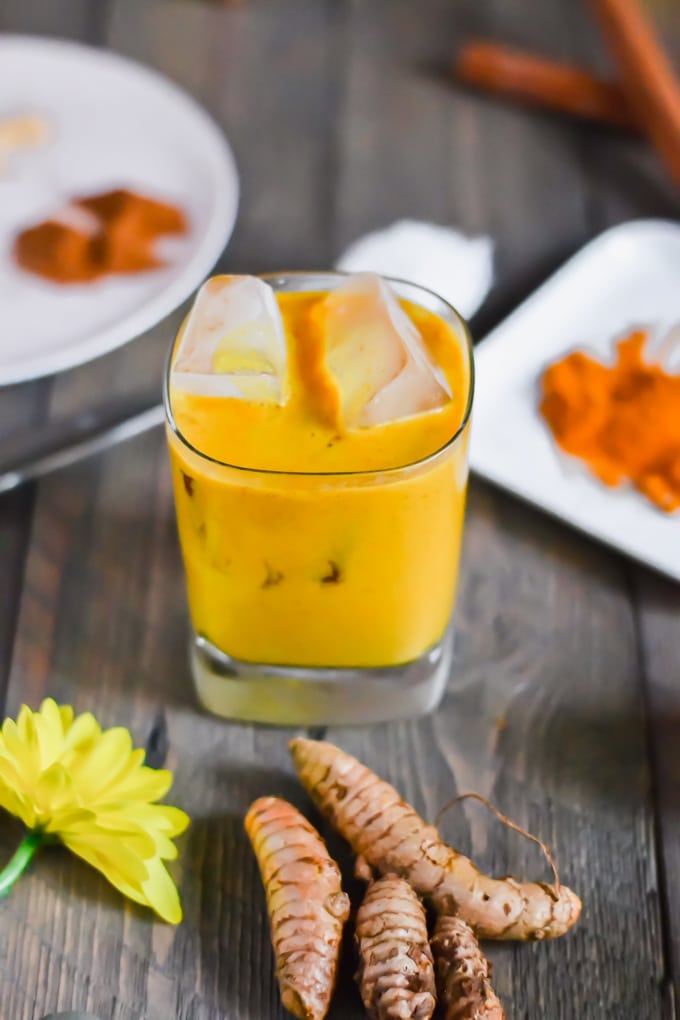 A glass with iced golden milk and fresh turmeric in the foreground.