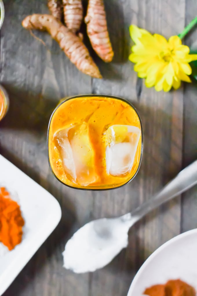 A glass of golden milk with ice cubes with a spoon of coconut oil and fresh turmeric in the background.