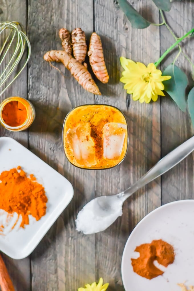 An aerial view of a glass of iced golden milk with ice cubes, a spoonful of coconut oil, fresh turmeric, and a yellow flower. 