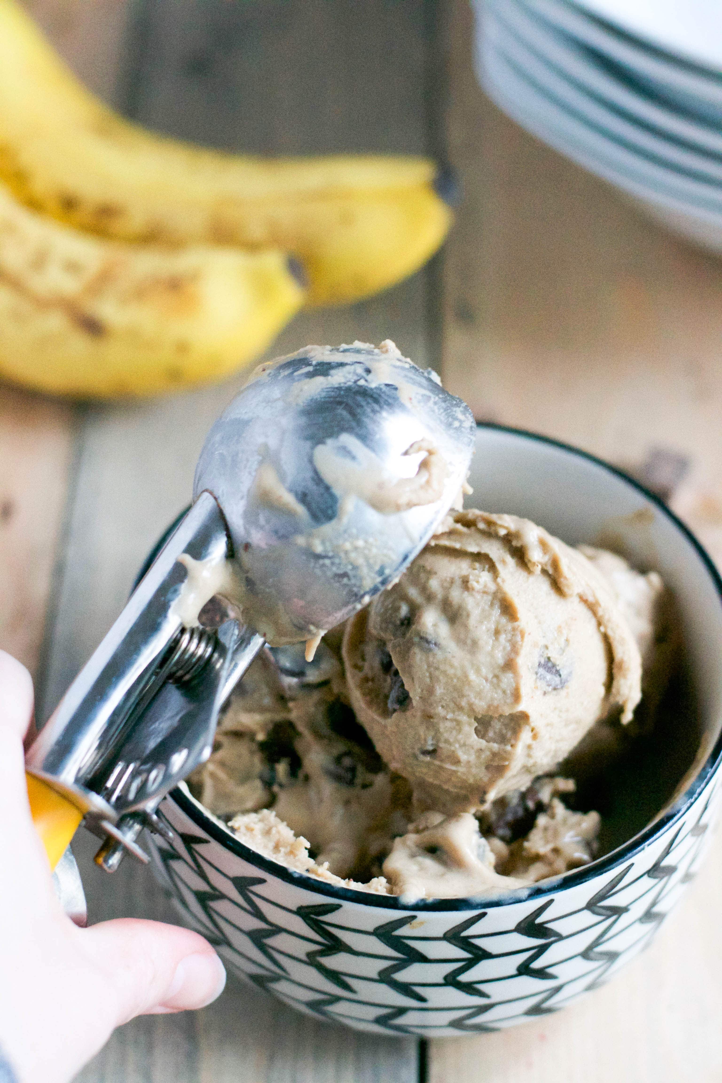An ice cream scooper placing a scoop of vegan banana ice cream with chocolate chunks into a bowl. 