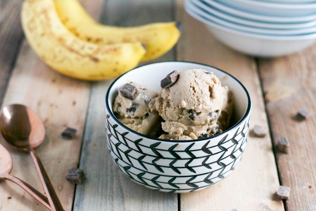 A bowl of vegan peanut butter banana ice cream with chocolate chunks next to spoons and a bunch of bananas. 