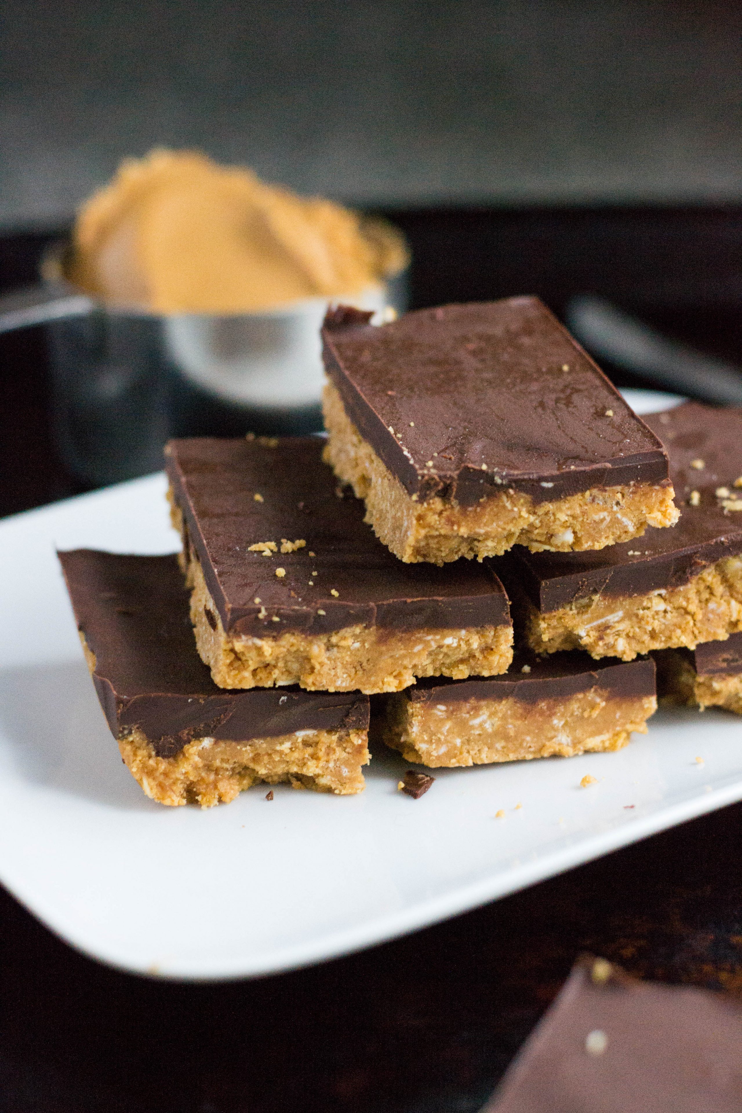 A pile of chocolate peanut butter bars on a white platter in front of a measuring cup of peanut butter. 