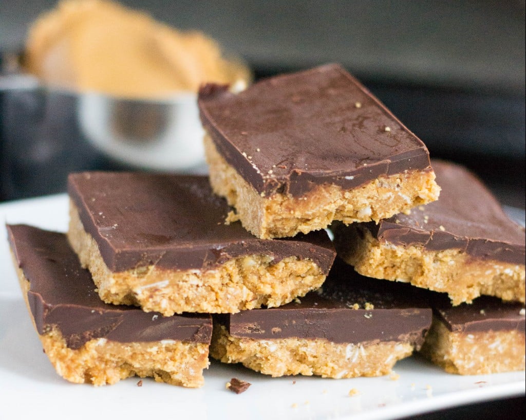 A pile of chocolate peanut butter bars in front of a measuring cup of peanut butter. 