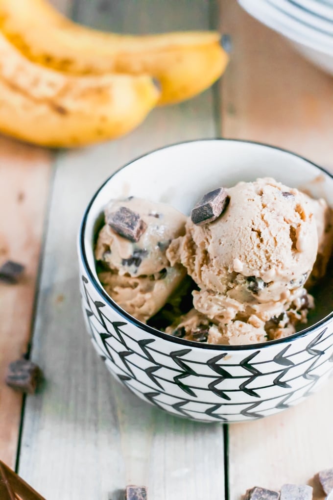 A bowl of vegan banana ice cream with chocolate chunks in front of a bunch of ripe bananas. 