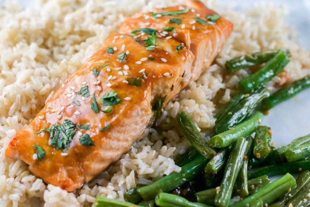 A portion of teriyaki salmon on top of a bed of brown rice with a side of green beans.