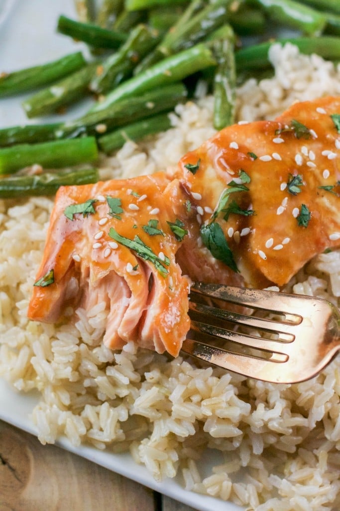 A plate of brown rice topped with green beans and teriyaki salmon with sesame seeds and a fork breaking into the salmon.