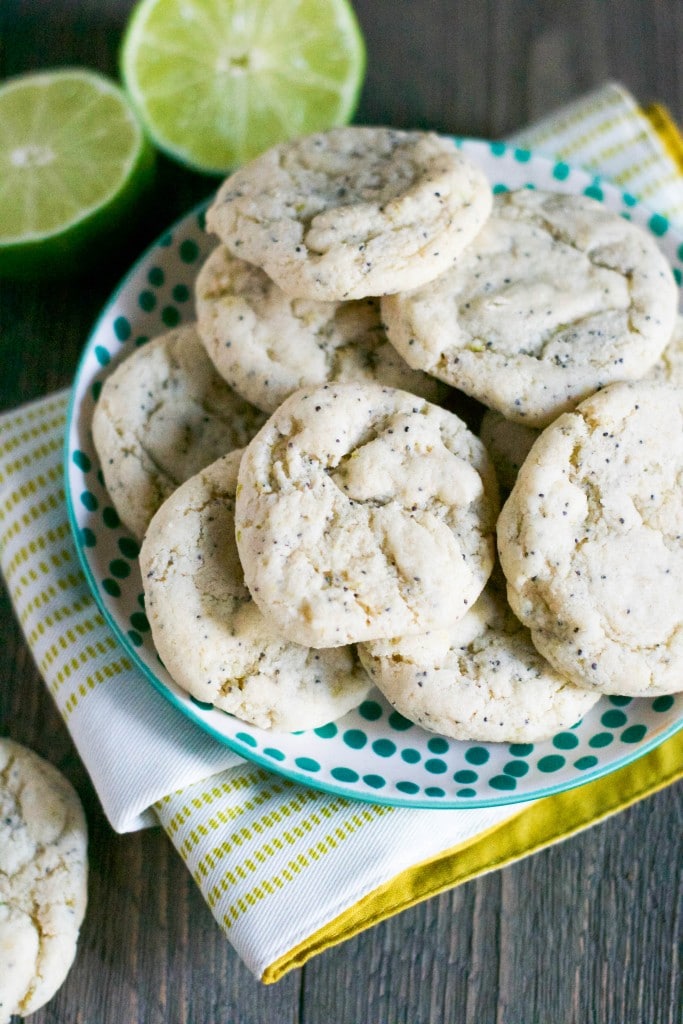 A pile of Gluten-Free Vegan Lime Poppyseed Cookies on a plate with lime halves on a table.