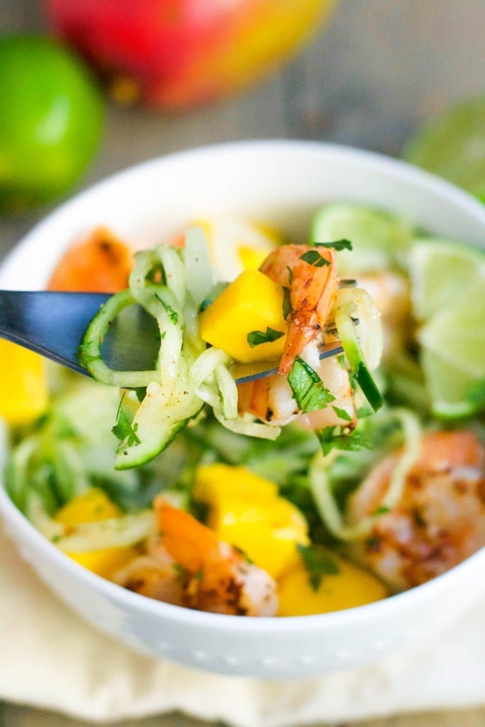 A fork with cucumber noodles, mango, shrimp and cilantro over a bowl of cucumber noodles. 