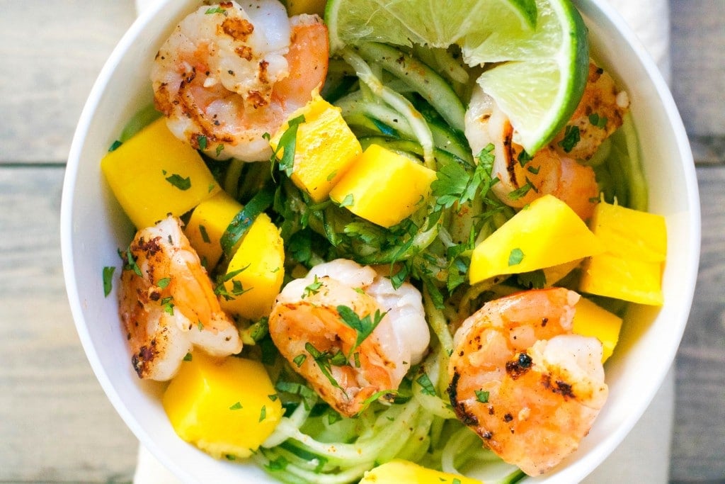 A bowl of cucumber noodles topped with mango, shrimp, cilantro and a lime wedge. 