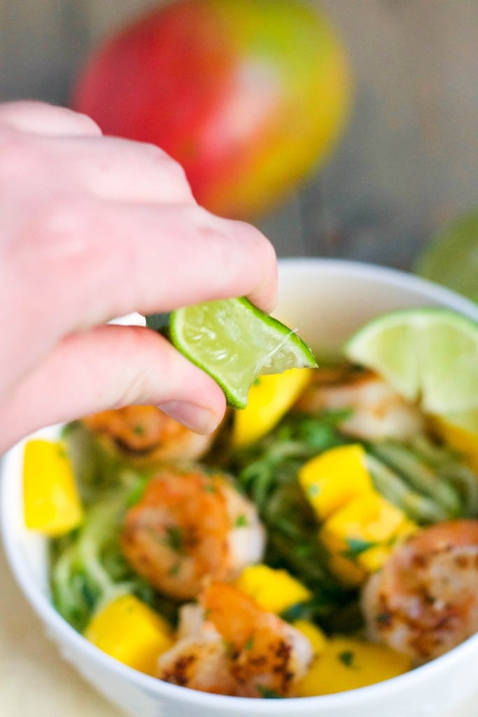 A hand squeezing lime over a bowl of cucumber noodles with shrimp and mango. 