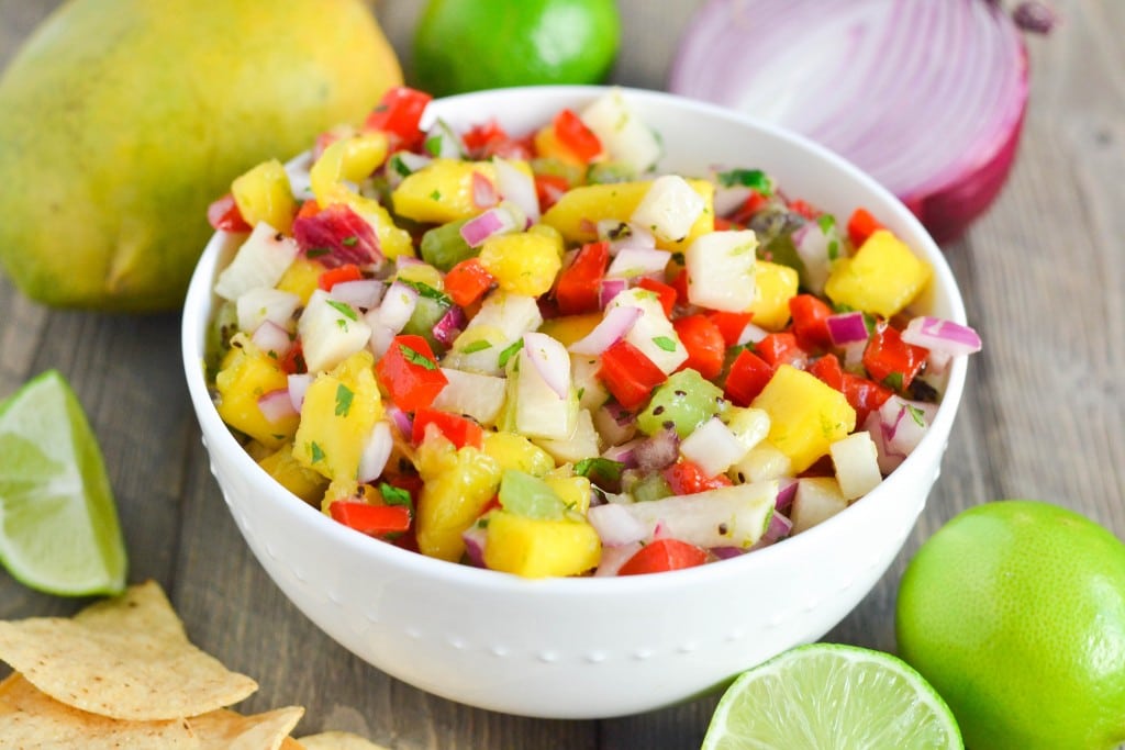 A bowl of kiwi mango jicama salsa with mangos, limes, tortilla chips and red onion in the background.