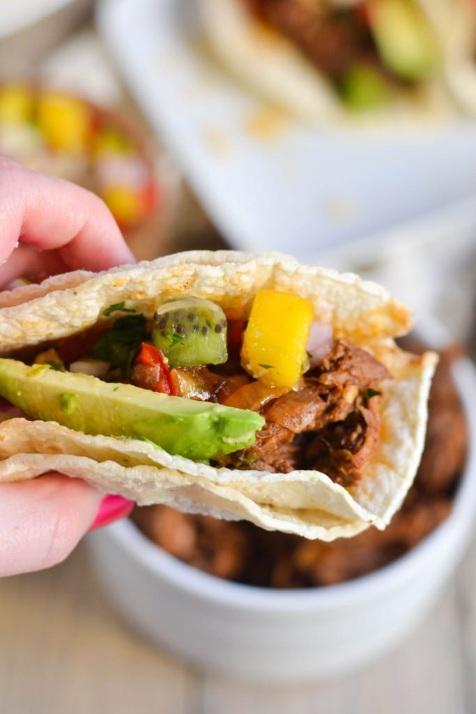 A hand holding a shredded beef taco with diced avocado, mango and kiwi salsa. 