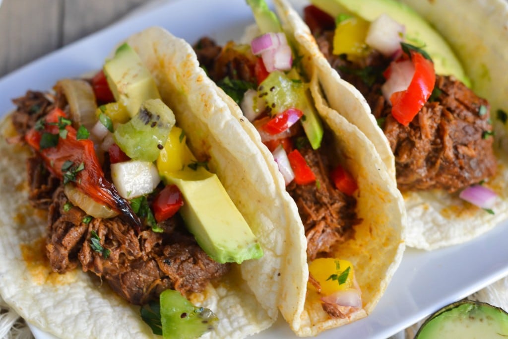 A platter of three Crock Pot Shredded Beef Tacos  topped with avocado, roasted peppers and kiwi. 