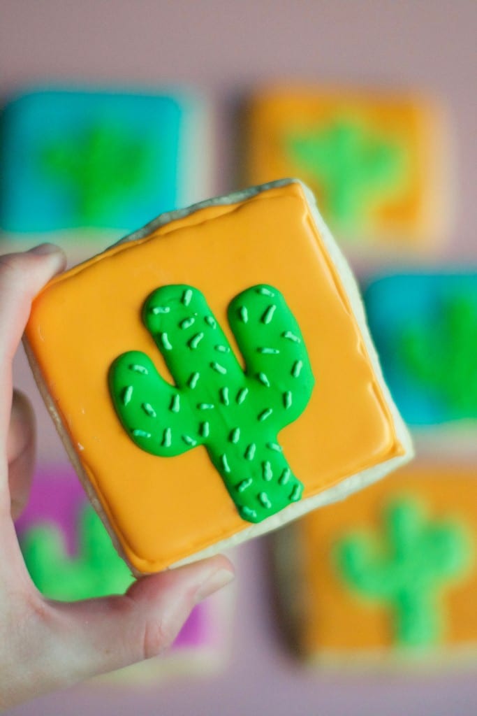 A hand holding a square sugar cookie decorated with royal icing to look like a cactus.