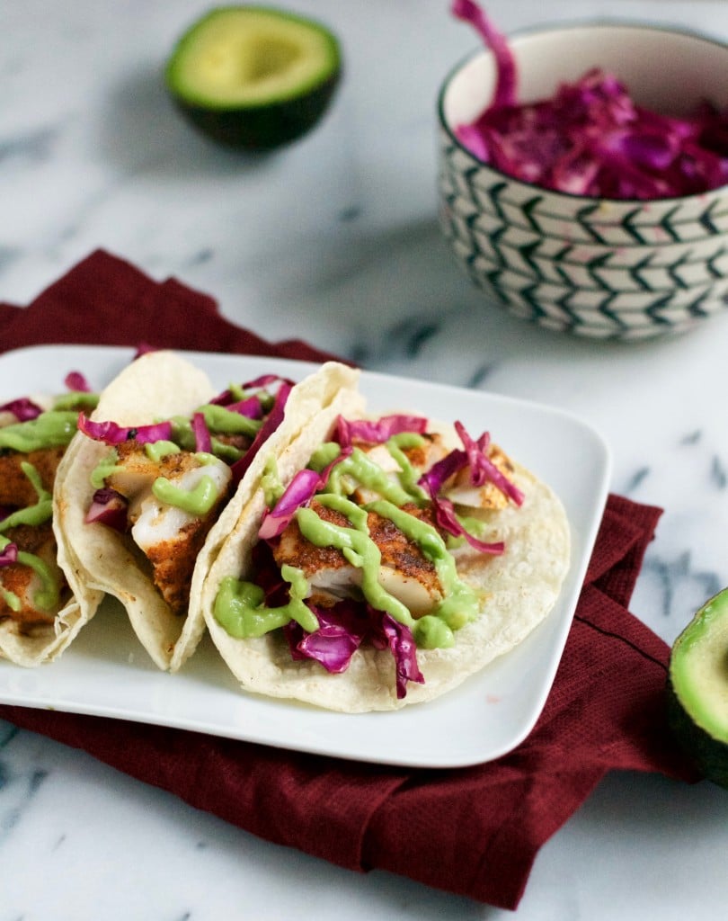 A plate with three blacked fish tacos topped with red cabbage and avocado crema in front of a bowl of red cabbage.