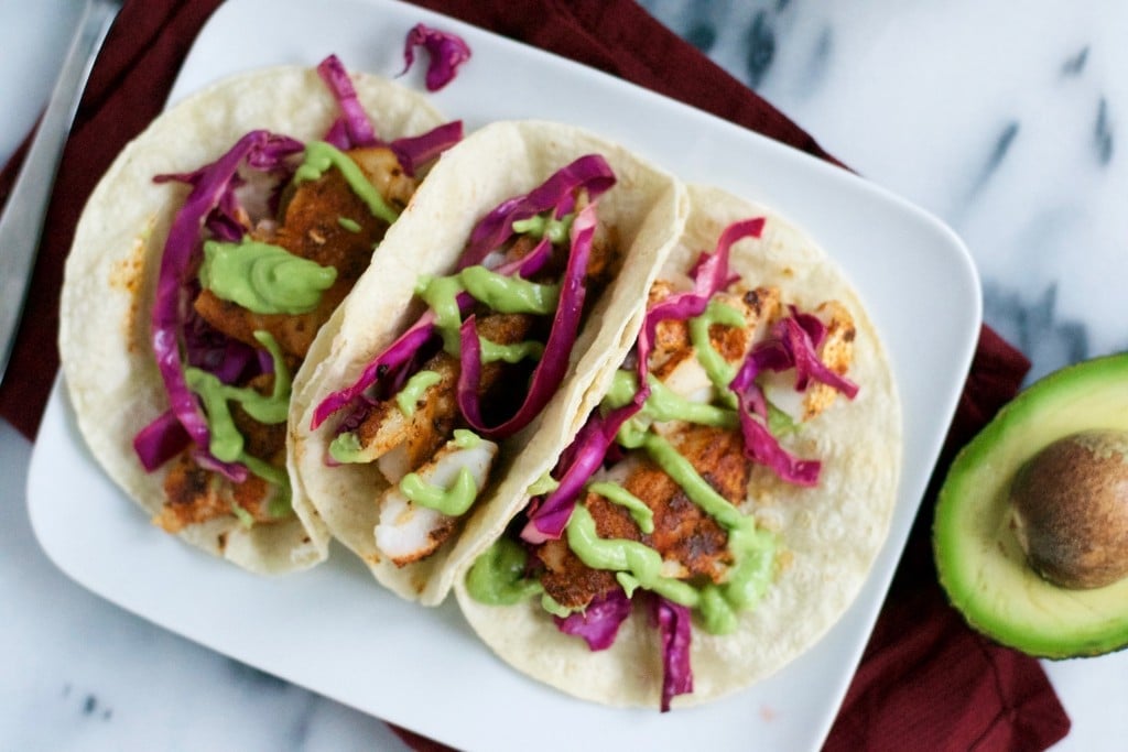 A platter of blackened fish tacos topped with red cabbage and avocado crema with a sliced avocado on the surface.