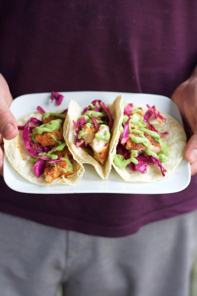 A man holding a plate with three blackened fish tacos with red cabbage and avocado crema.