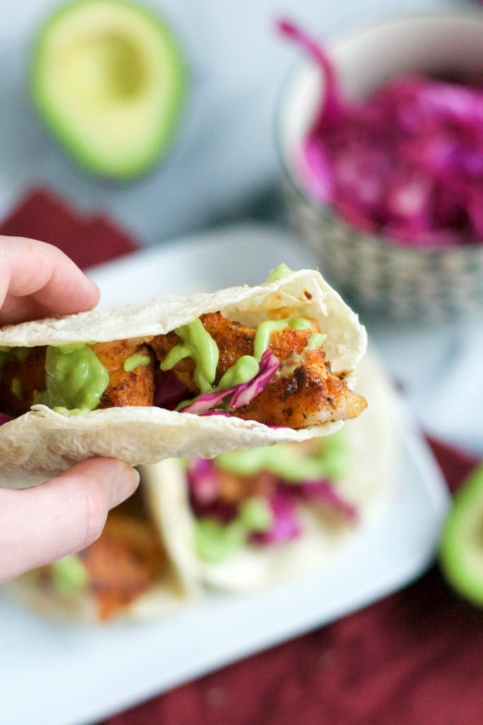 A hand holding a blackened fish taco topped with red cabbage and avocado crema. 