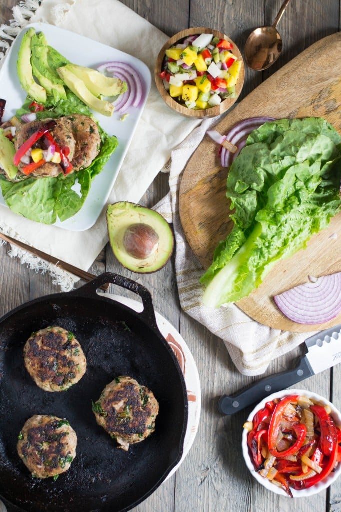 A skillet with garlic spinach turkey burgers, a cutting board with lettuce, a bowl of peppers, and a side of mango salsa.