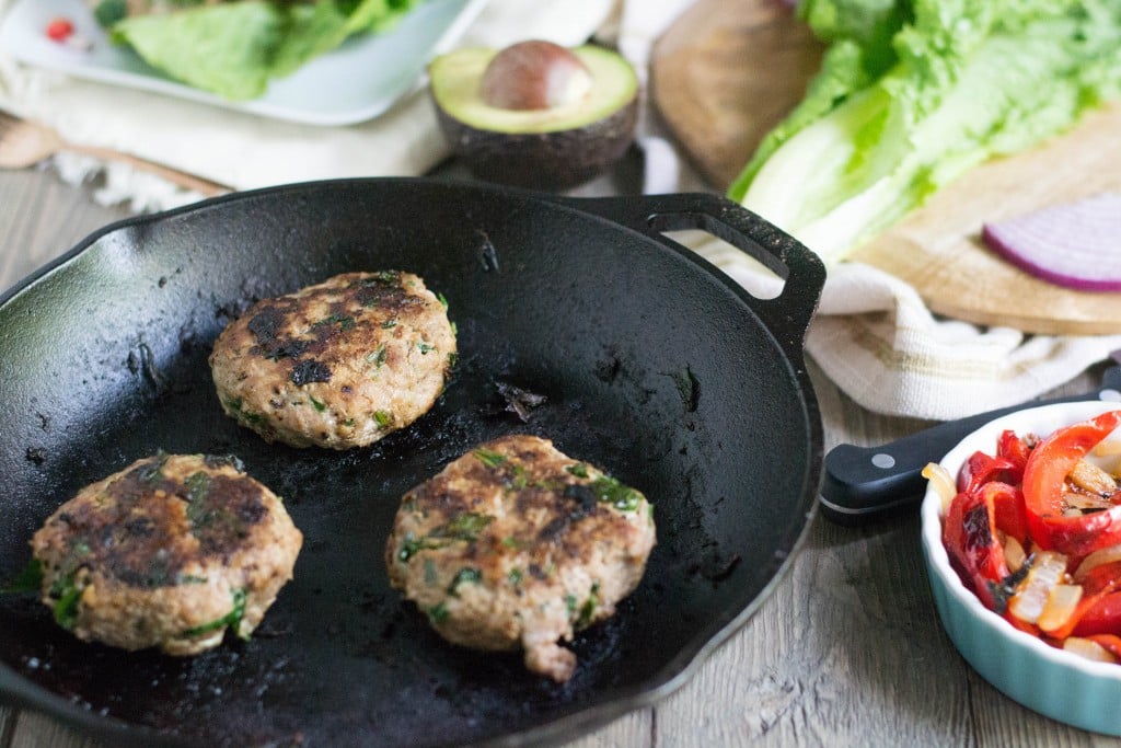 A cast iron skillet with three Spinach Garlic Turkey Burgers a side of roasted peppers and lettuce on a cutting board.