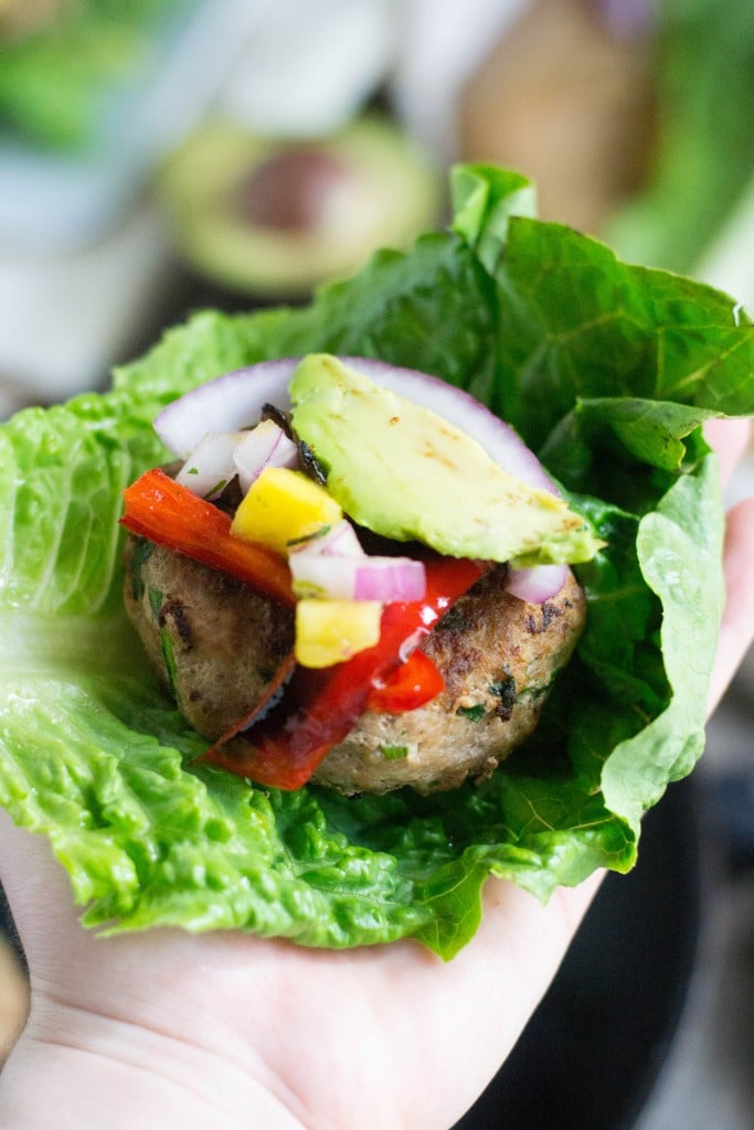 A hand holding a lettuce wrap with a spinach garlic turkey burger topped with sliced avocado, roasted peppers and mango salsa. 