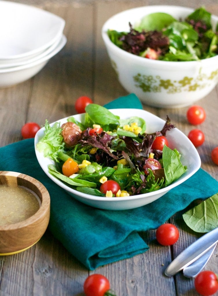 A bowl of salad with tomato, corn, green beans and potatoes with a bowl of honey mustard dressing and cherry tomatoes scattered on the surface.